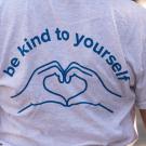 The back of a student wearing a light gray T-shirt with a blue linear outline of two hands cupped to form a heart and above the words "Be Kind to Yourself."