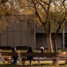 Three students sit on a bench facing a two-story concrete building. They wear cold weather clothing and the surrounding trees are bare from the winter season.