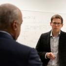 Two men converse in a bright room with whiteboards covered in notes and diagrams.