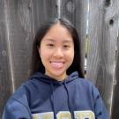 Roslyn stands in front of a gray wooden fence. She wears her log black hair down behind her shoulders and a dark blue hoodie sweatshirt that has the yellow letters UCD on the front and in a blue strip in the middle of them running the length of the chest in yellow letters it says University of California, Davis.