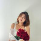 Monica sits in front of a white wall with her left knee bent up in front of her. She wears her dark brown shoulder length hair down straight in a bob, a gold necklace with a circular charm, a white tank top and blue jeans ripped at the knee. She holds a bouquet of a dozen red roses wrapped in a white florist paper across her body. 