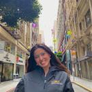 Ellie stands in a narrow street with tall buildings on either side and round paper lanterns in bright colors strung overhead. Her long straight dark brown hair falls behind her shoulders and she wears a navy zip-up jacket with the words UC Davis Veterans Success Center on the right chest.