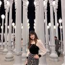 Ashley stands illuminated in the night by dozens of ornate lampposts crowded together in the space and in the background. She smiles and stands posed in front of the lampposts with has her left hand on her hip. Her long straight black hair is styled straight and with bangs and it falls in front of her shoulders. She wears a black top with white sheer sleeves and black cuffs and a short black and white tweed wrap skirt with three gold buttons running down the front.