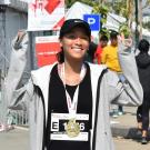 Brooke stands with her arms raised to the height of her head as she celebrates completing a marathon. She wears a runners bib pinned to her black top under an open light gray zip-up hoodie and a marathon brass metal in the shape of the number 6 around her neck on a white ribbon. Her black hair is worn in a ponytail under a black hat with a white Nike swoosh on the front.