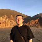 Yuchen stands in the setting sunlight on medium brown dirt that resembles wet sand littered with footprints and tracks. Behind him are rounded hills of dirt set against a clear blue sky. His black hair is buzzed and he wears round metal-framed glasses, a black T-shirt and a thick black nylon bag strap across his body.