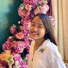 Asuka has long straight black hair and wears a white track jacket with the word AIR and a Nike swoosh stitched in black on the left side. She sits at a table in doors with a small bud vase filled with delicate florals in front of her and with an abundance of pink flowers cascading down behind her on a deep green wall with a cream colored drape.