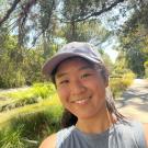 Amy wears a light gray hat and gray sleeveless shirt in a selfie taken in the daylight at the Arboretum at UC Davis with plants, trees and a wooden bridge in the distance behind her.