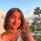 Selfie of Sana overlooking palm trees and cream colored hotel or apartment buildings. She is cast in light from the setting sun and wears a white sleeveless top with vertical black stripes, a silver necklace with an intricate charm, and her bard brown hair is a little longer than chin length worn in a middle part and straight down. The top of her palm tree tattoo is just visible on her left shoulder.