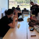 Six students around a table with their instructor working on a laptop at the front of the room. One student in a UC Davis T shirt stands and gives a peace sign with two fingers to the camera.