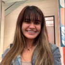 Trisha has long brown hair and bangs. She stands inside for a selfie. She wears a medium-wash jean jacket over a taupe top, and has a gold necklace with the letter T. 