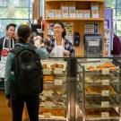 Inside the UC Davis Coffee House, a student worker in a backward hat smiles as they extend their arm with a pasty bag in their hand over a tall counter to a student customer on the other side who wears a backpack.