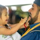 A man with a dark beard wears a cap and gown while holding a toddler girl in a white dress and a white bow in her dark brown hair. She holds her hands up to his face and smiles at him as he smiles at her playfully in the middle of their game.