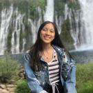 Jackie sits on a rock with water falls in the background pouring down a sheer rockface with greenery climbing up the face of the cliff and pouring into a large pool of water. Jackie's long straight black hair is worn long. She wears a destressed medium blue jean jacket over a white tank top with black, teal and coral striping and black shorts with a white draw string.