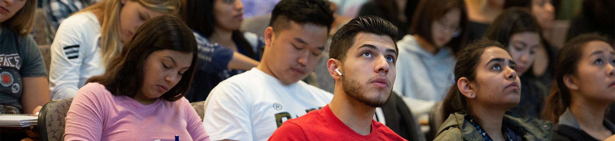 A large group of students seated in a lecture hall take notes and look toward the front of the room.