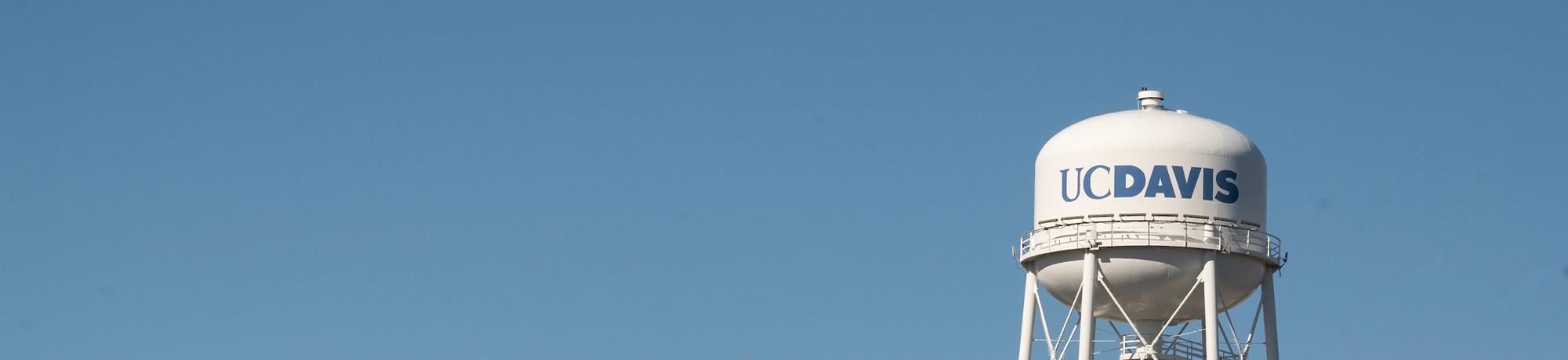 The UC Davis logo in blue on a large white water tower against a clear blue sky.