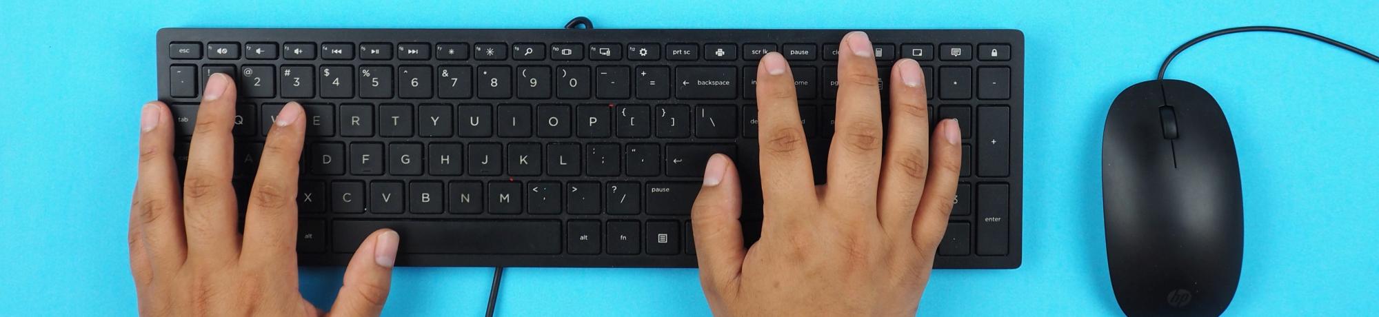 hands on a black computer keyboard with a black mouse to the right. The keyboard and mouse sit on a bright blue table top.