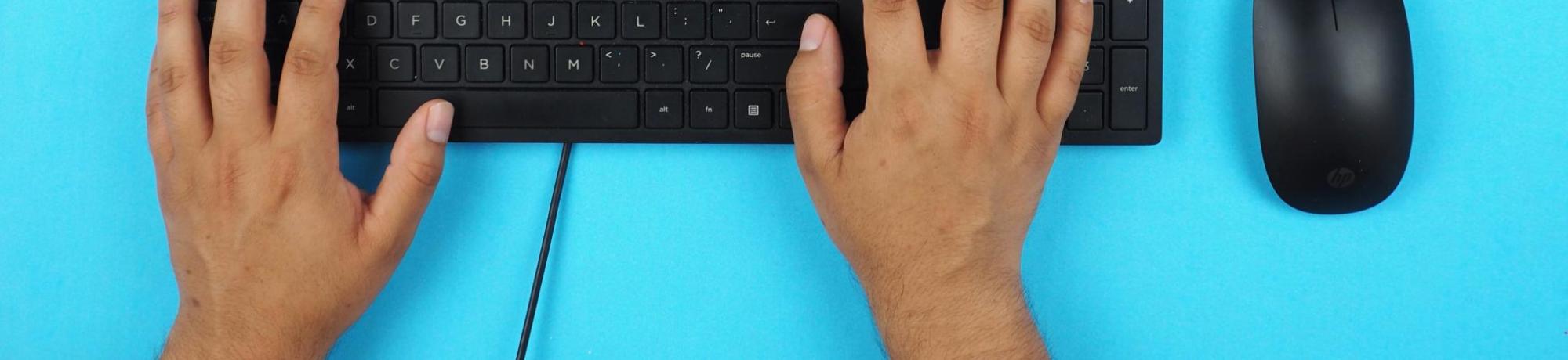 hands on a black computer keyboard with a black mouse to the right. The keyboard and mouse sit on a bright blue table top.