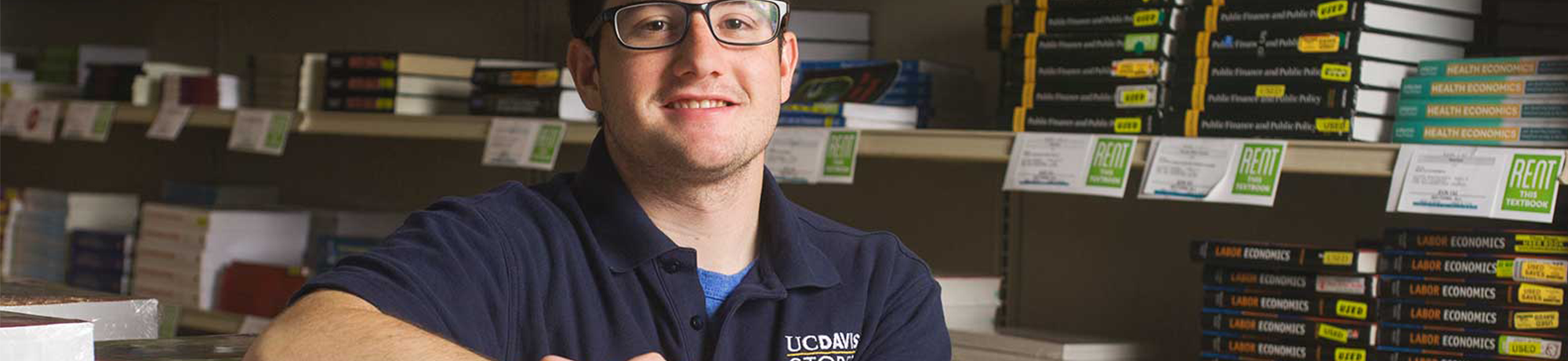 A student smiles at the camera with his arm propped up on a stack of books in a room with filled book shelves. His polo shirt reads "UC Davis Stores."